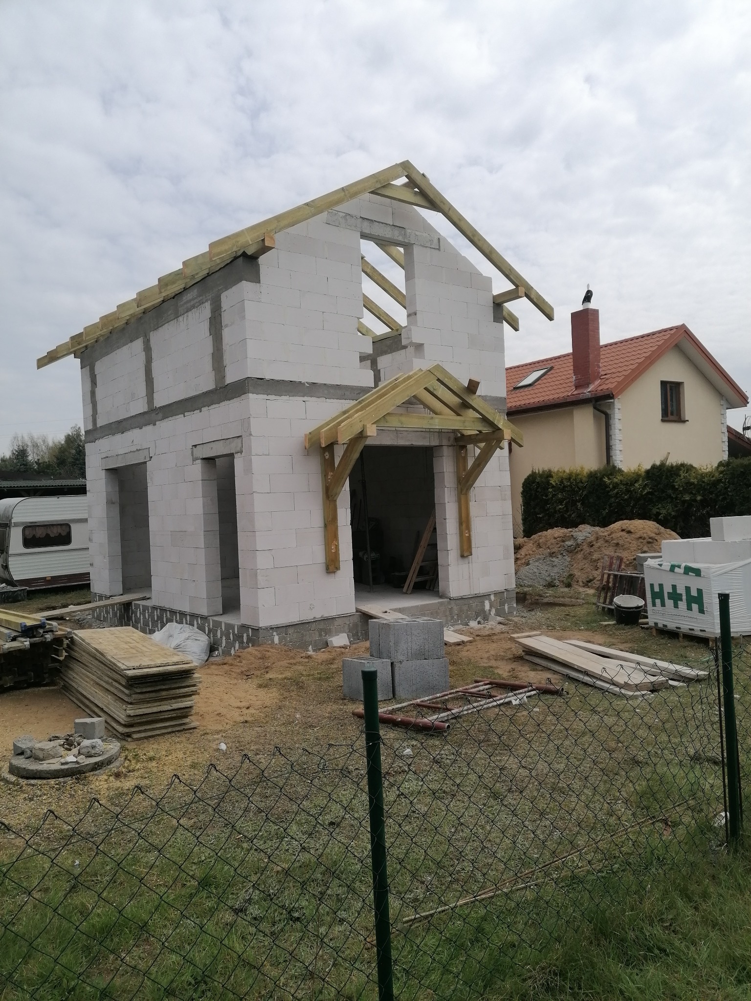 Surowy stan domu z białych bloczków, widoczne drewniane elementy konstrukcji dachu i nadproży, na pierwszym planie siatka ogrodzeniowa, w tle dom z czerwoną dachówką.