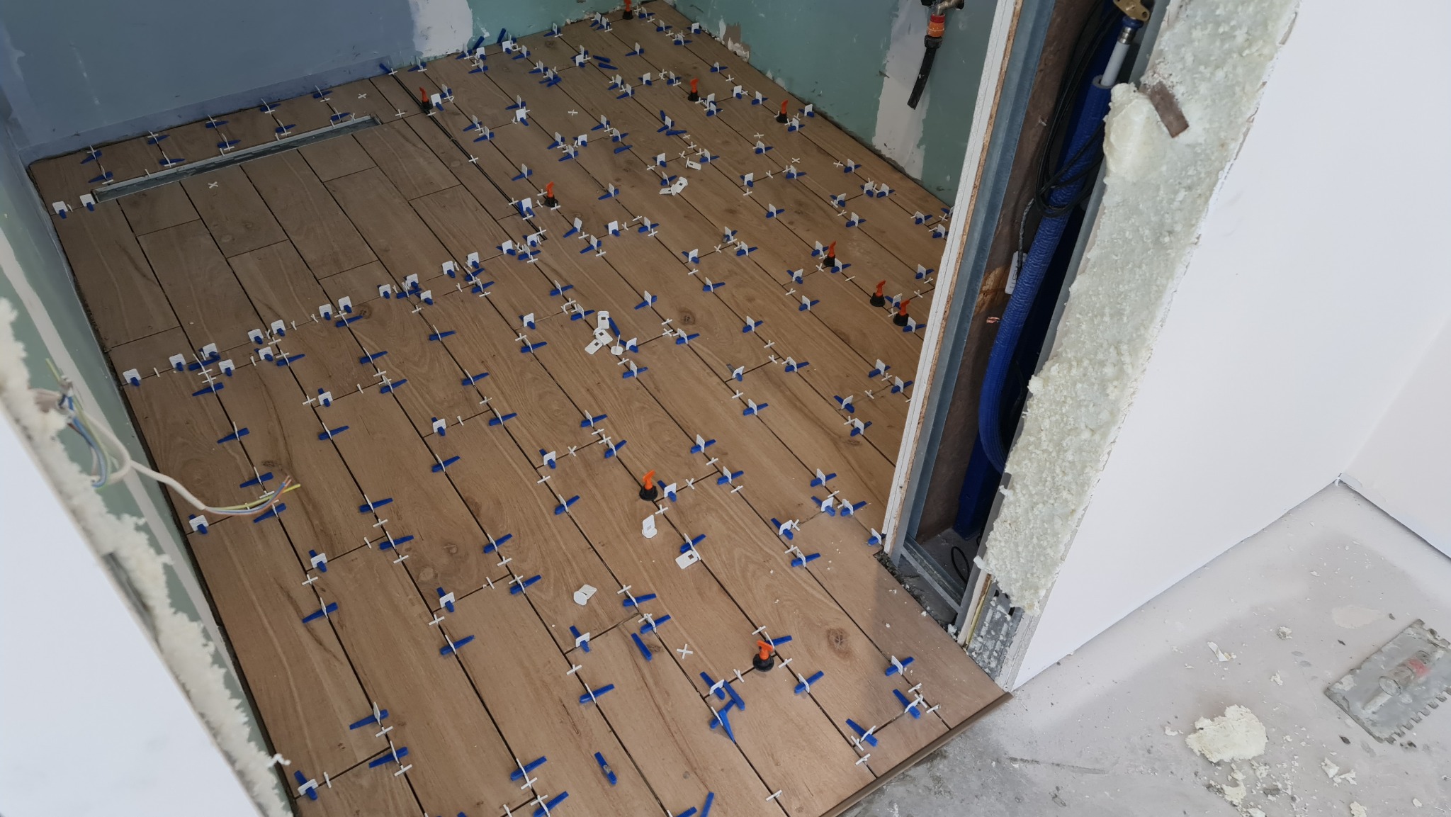 Układanie brązowych płytek imitujących drewno w łazience, z zastosowaniem systemu poziomowania płytek z niebieskimi krzyżykami i pomarańczowymi klinami. Widoczny odpływ liniowy w podłodze...