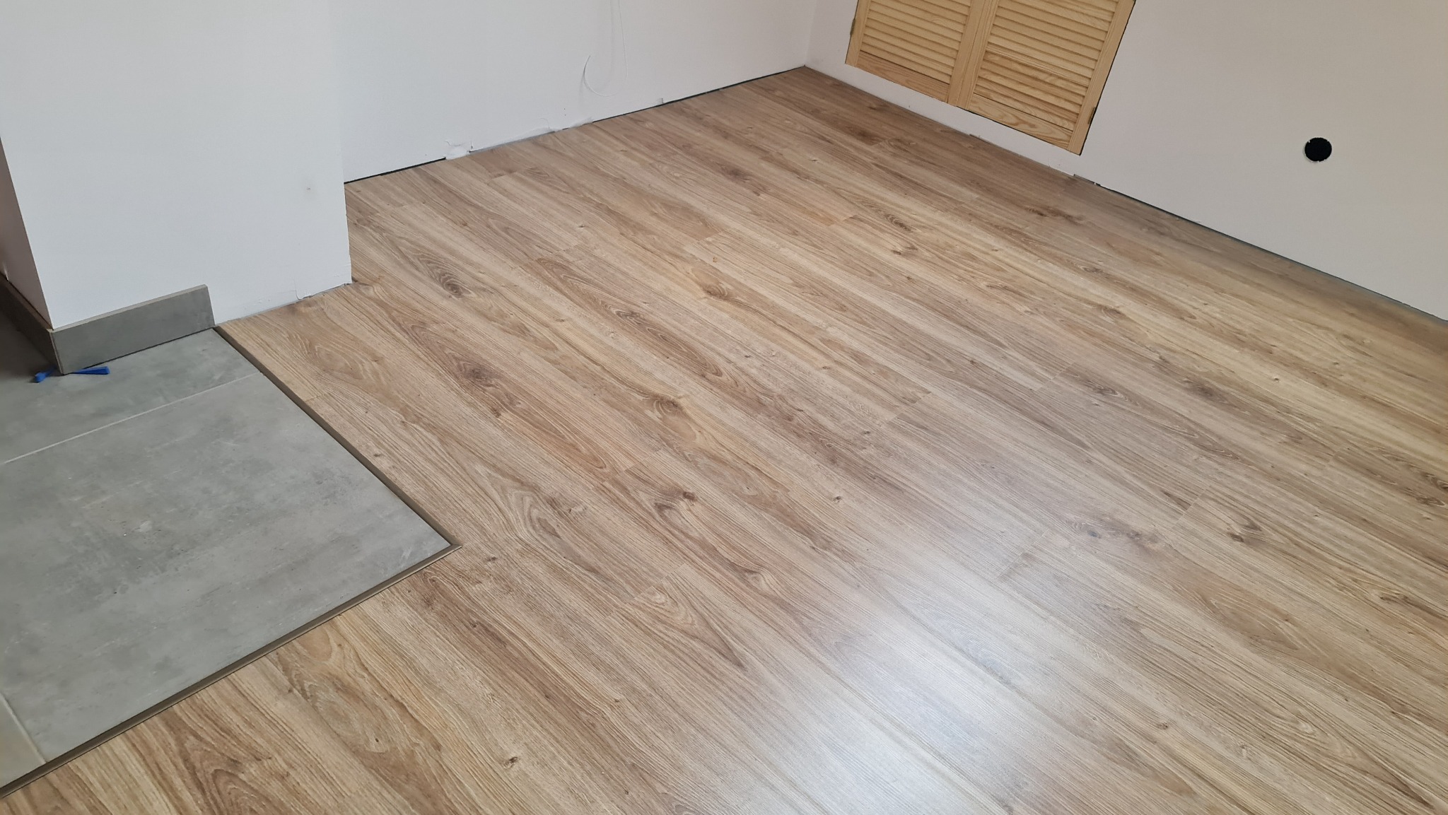 Jasne panele podłogowe ułożone w nowym pomieszczeniu, z fragmentem szarej podłogi wyłożonej płytkami i listwą przypodłogową. Widoczne narzędzie do układania paneli.