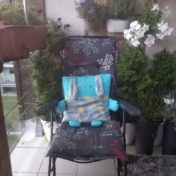 Na balkonie, na krześle z poduszką w kwiaty leży turkusowa poduszka w kształcie królika z szarą kamizelką w kolorowe kółka.