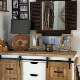 Karolina Gołębiewska Oliart - Rustykalna komoda z przesuwanymi drzwiami i białymi szufladami, dekoracje w postaci drewnianej lampy, ramek ze zdjęciami i lustra z okiennicami.