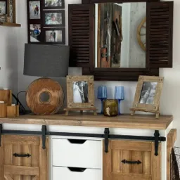 Rustykalna komoda z przesuwanymi drzwiami i białymi szufladami, dekoracje w postaci drewnianej lampy, ramek ze zdjęciami i lustra z okiennicami.