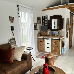 Karolina Gołębiewska Oliart - Salon w stylu rustykalnym z drewnianą szafką z przesuwanymi drzwiami, białymi frontami szuflad, lustrem w drewnianej ramie i dekoracjami. Na pierwszym planie stolik kawowy z czerwonym imbrykiem...