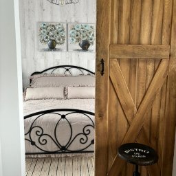 Karolina Gołębiewska Oliart - Rustykalne drzwi przesuwne z drewna, widok na sypialnię z metalowym łóżkiem i dekoracjami ściennymi, stołek z napisem 'Bistro de Paris'.