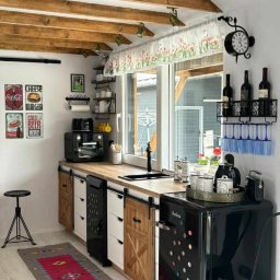 Karolina Gołębiewska Oliart - Retro kuchnia z drewnianymi belkami stropowymi, przesuwnymi frontami szafek w stylu rustykalnym, czarną lodówką retro i dywanem w etniczne wzory.