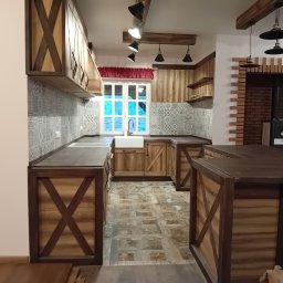 Karolina Gołębiewska Oliart - Rustykalna aranżacja wnętrza: drewniane szafki z krzyżowymi zdobieniami, biała ceramiczna umywalka pod oknem, wzorzyste płytki na ścianie i podłodze, schody z boku.