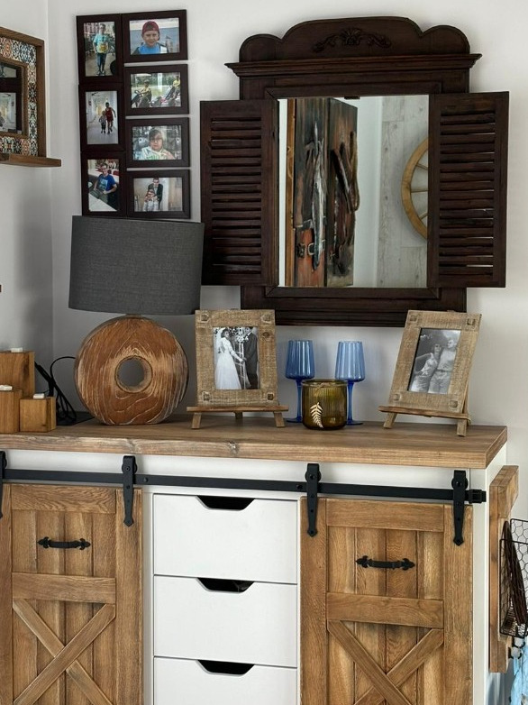 Rustykalna komoda z przesuwanymi drzwiami i białymi szufladami, dekoracje w postaci drewnianej lampy, ramek ze zdjęciami i lustra z okiennicami.