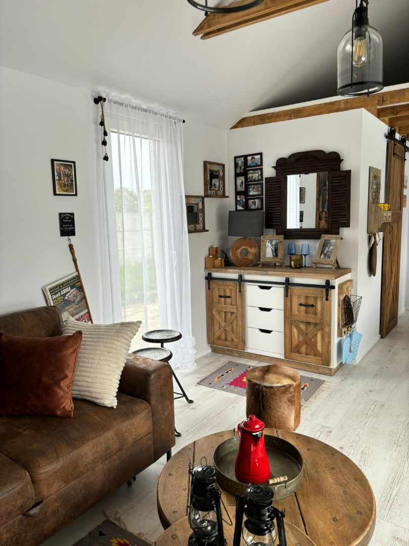 Salon w stylu rustykalnym z drewnianą szafką z przesuwanymi drzwiami, białymi frontami szuflad, lustrem w drewnianej ramie i dekoracjami. Na pierwszym planie stolik kawowy z czerwonym imbrykiem...