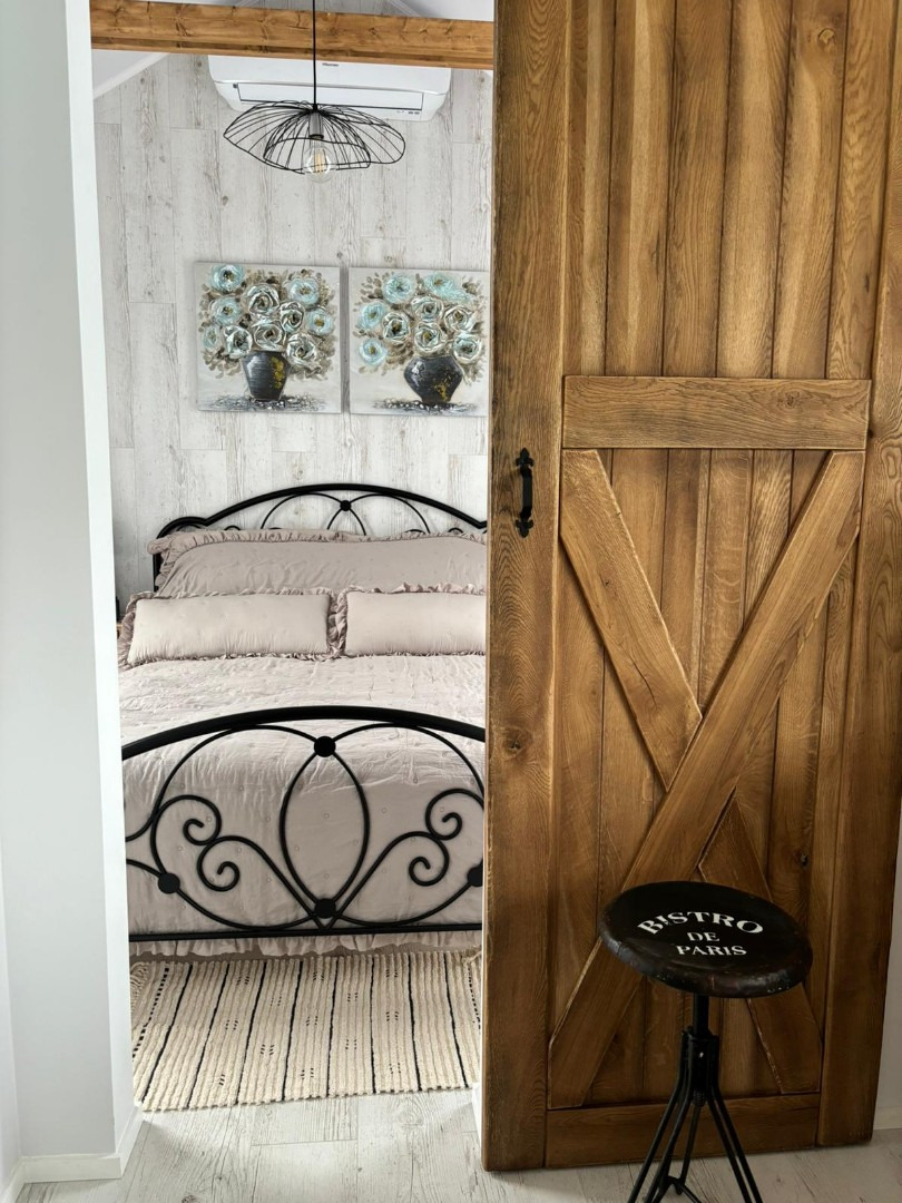 Rustykalne drzwi przesuwne z drewna, widok na sypialnię z metalowym łóżkiem i dekoracjami ściennymi, stołek z napisem 'Bistro de Paris'.