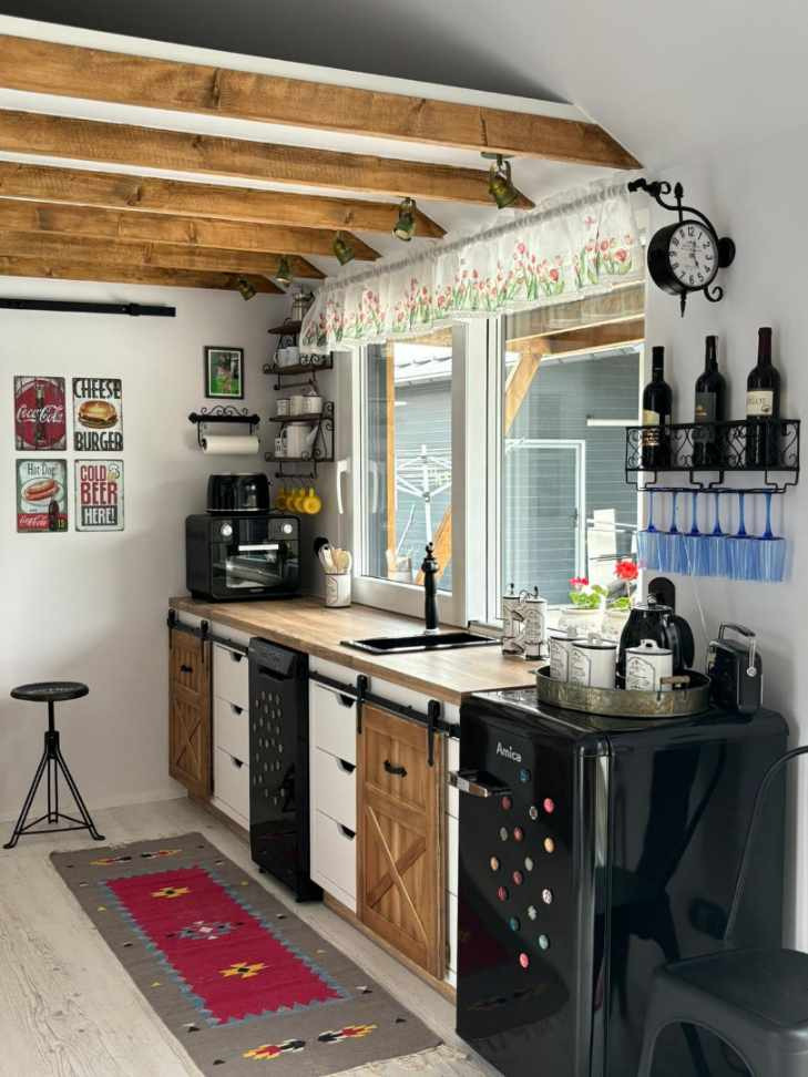 Retro kuchnia z drewnianymi belkami stropowymi, przesuwnymi frontami szafek w stylu rustykalnym, czarną lodówką retro i dywanem w etniczne wzory.