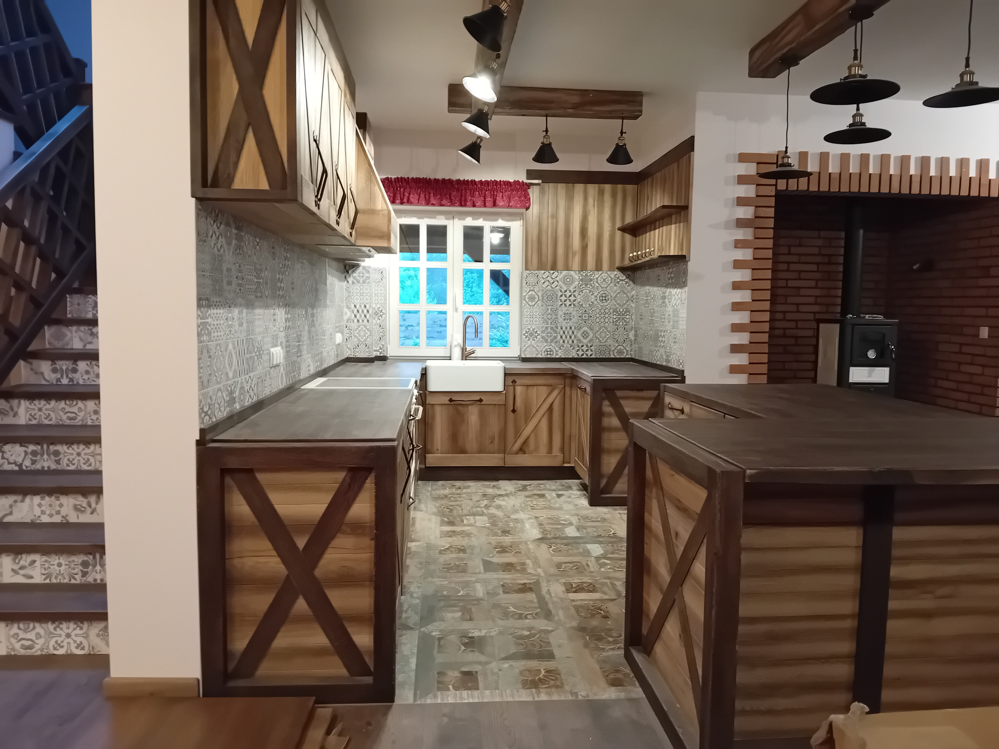 Rustykalna aranżacja wnętrza: drewniane szafki z krzyżowymi zdobieniami, biała ceramiczna umywalka pod oknem, wzorzyste płytki na ścianie i podłodze, schody z boku.