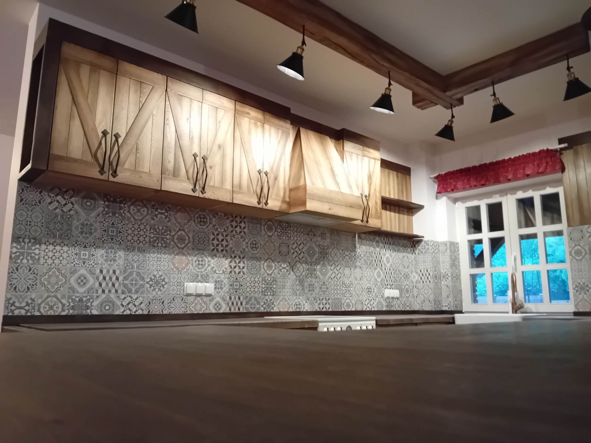 Drewniane szafki kuchenne z rustykalnymi uchwytami i wzorzystymi płytkami nad blatem w kuchni w Wilczu, oświetlone wiszącymi lampami.