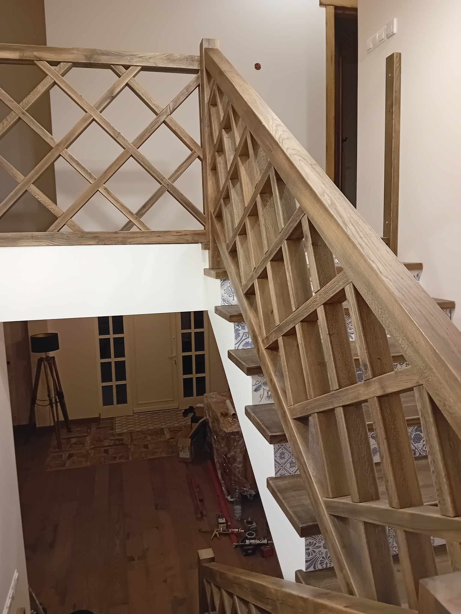 Drewniane schody z balustradą o kratowym wzorze, stopnie wykończone płytkami z delikatnym wzorem, widok z góry na dół, w tle widoczne drzwi i lampa na trójnogu.