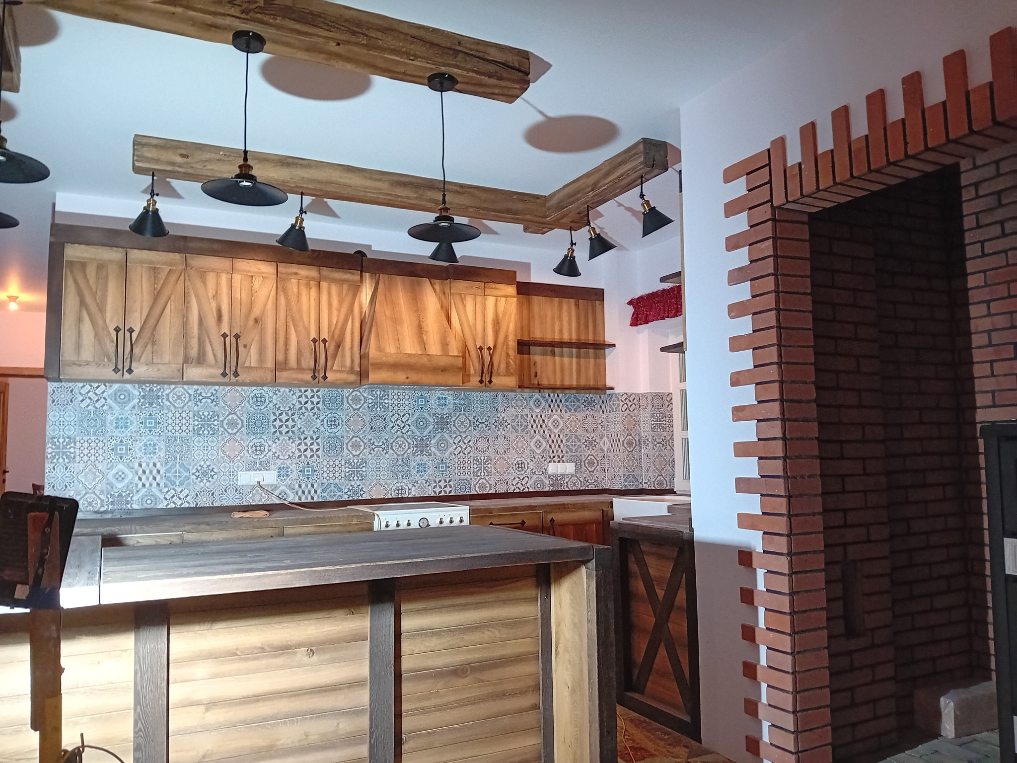 Rustykalna aranżacja: drewniane szafki z krzyżowymi zdobieniami, wzorzysta mozaika nad blatem, sufit z drewnianymi belkami i czarnymi lampami, ceglane obramowanie przejścia.