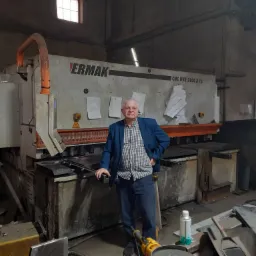 Mężczyzna w niebieskiej marynarce stoi przy maszynie CNC ERMAK HVR 3100 X 13 w warsztacie, obok leżą metalowe elementy.