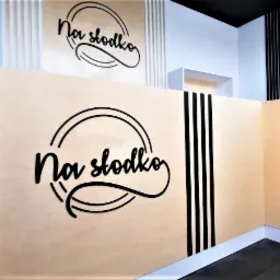 Drewniana zabudowa lady sklepowej z czarnymi paskami i logo 'Na słodko' w czarnej czcionce kursywą, widok z boku na jasne wnętrze sklepu.