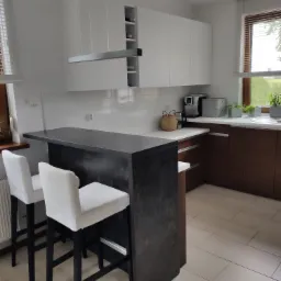 Nowoczesna kuchnia z białymi szafkami wiszącymi, drewnianymi szafkami dolnymi i ciemnym blatem wyspowym z dwoma białymi krzesłami barowymi.