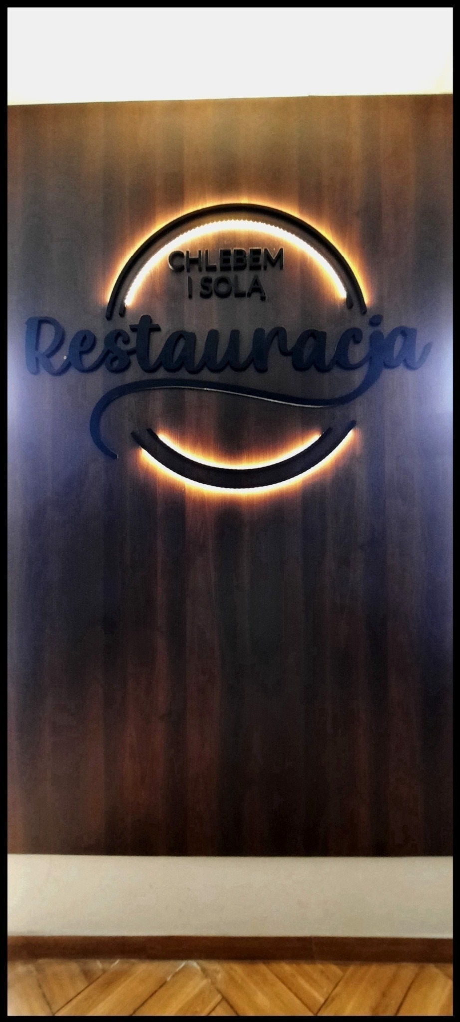 Podświetlany szyld restauracji 'Chlebem i Solą' na drewnianej ścianie.