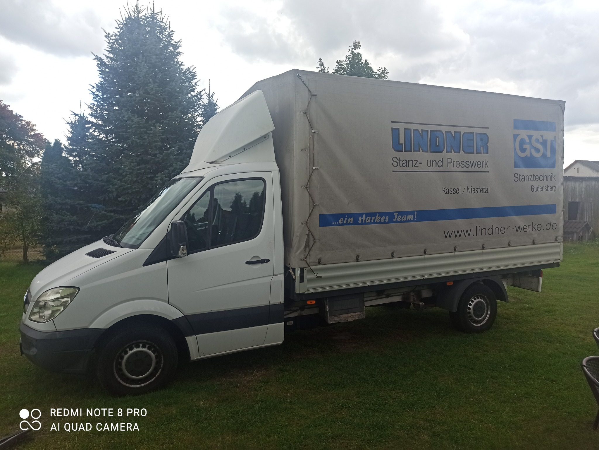 Biały samochód dostawczy z plandeką z logo firmy Lindner Stanz- und Presswerk, stojący na trawie przed świerkami.