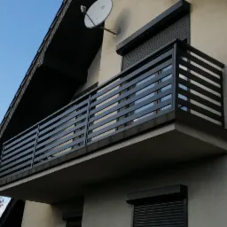 Nowoczesna, pozioma, czarna balustrada balkonowa z metalowymi profilami na tle beżowej elewacji domu i błękitnego nieba.