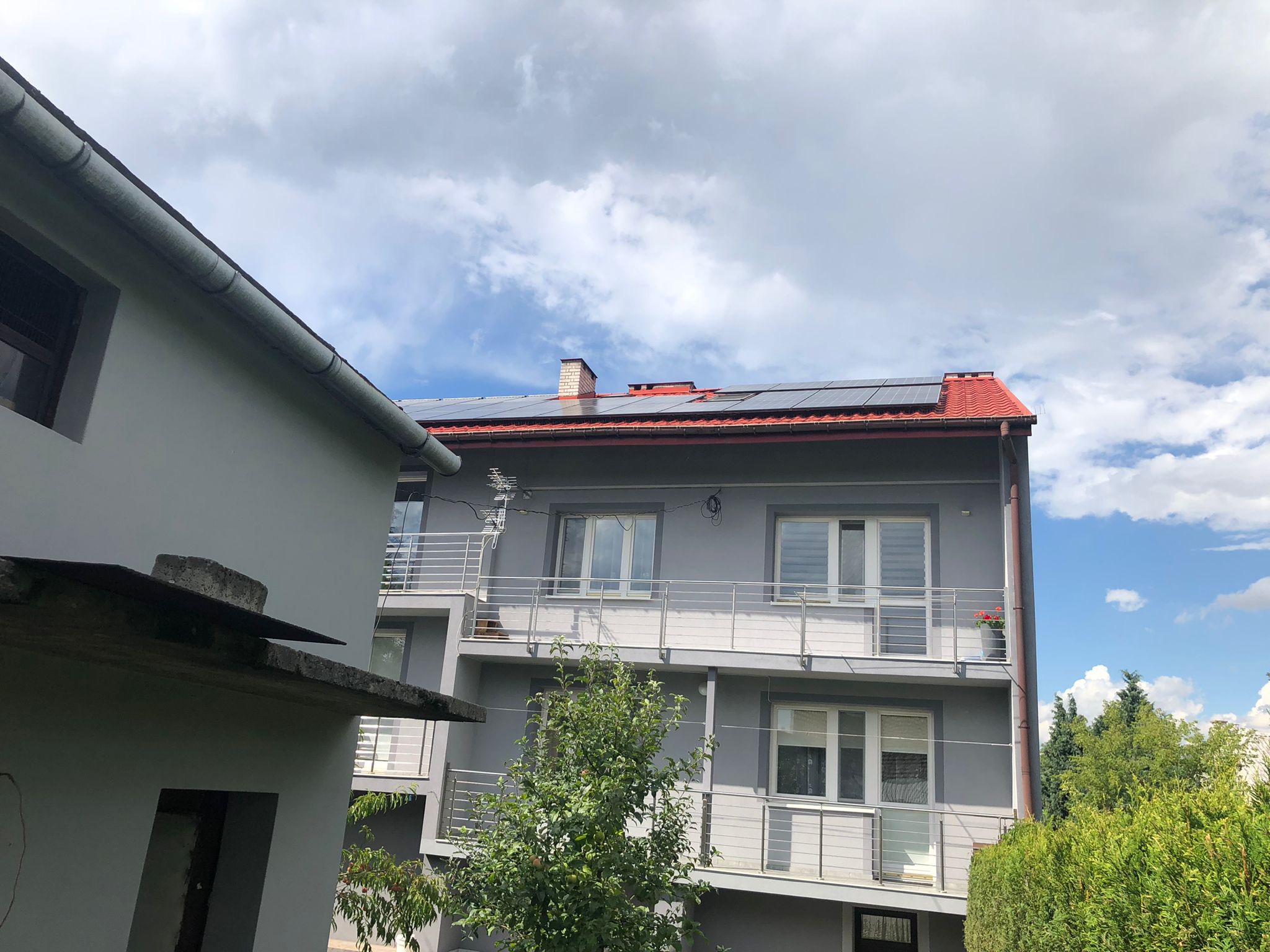 Dwupiętrowy szary dom z czerwonym dachem i panelami słonecznymi na dachu, widok z dołu na tle częściowo zachmurzonego nieba.