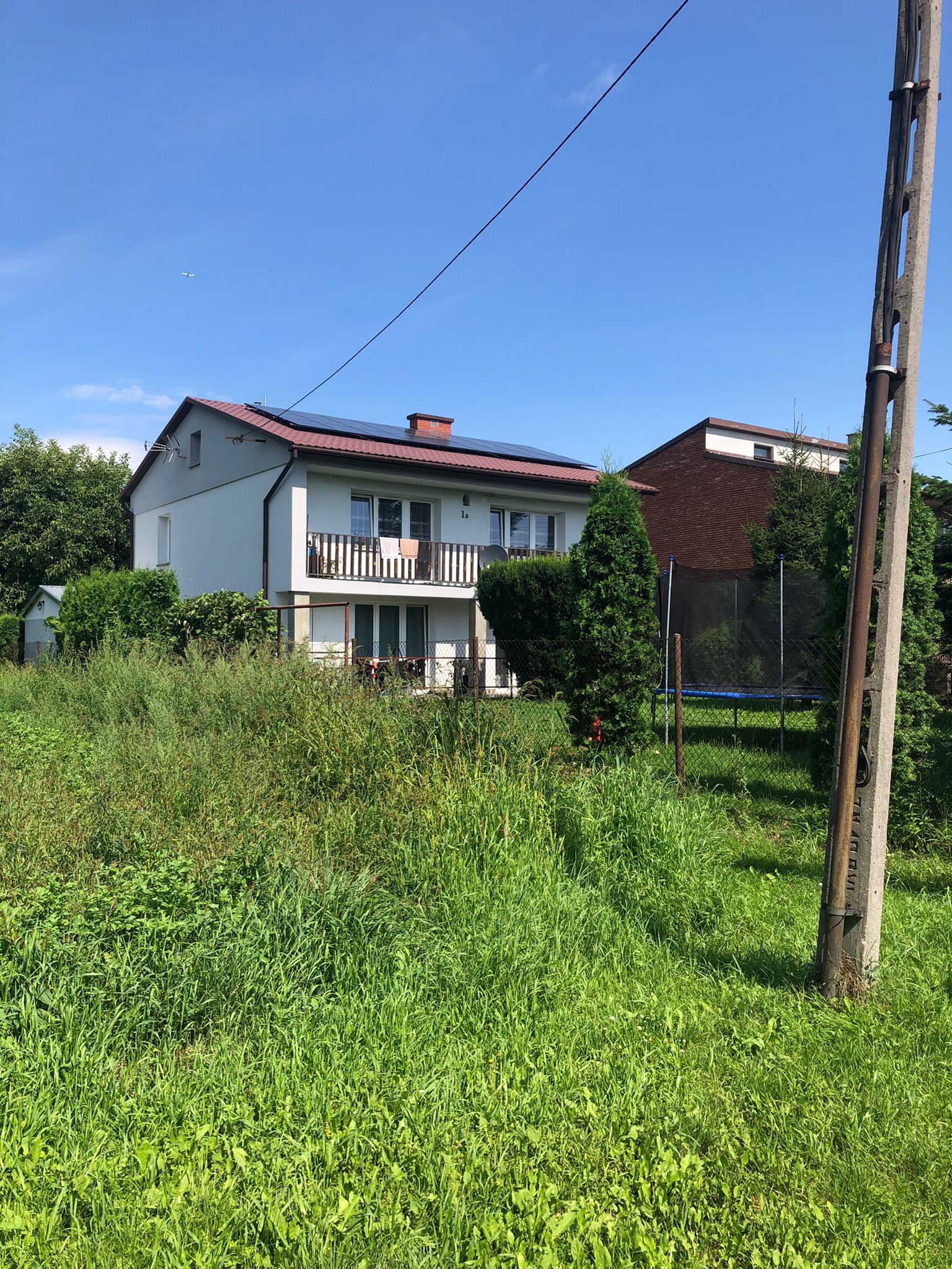 Dom jednorodzinny z panelami fotowoltaicznymi na dachu, widoczny zza zarośniętego trawnika i słupa energetycznego. Na balkonie suszą się ręczniki.