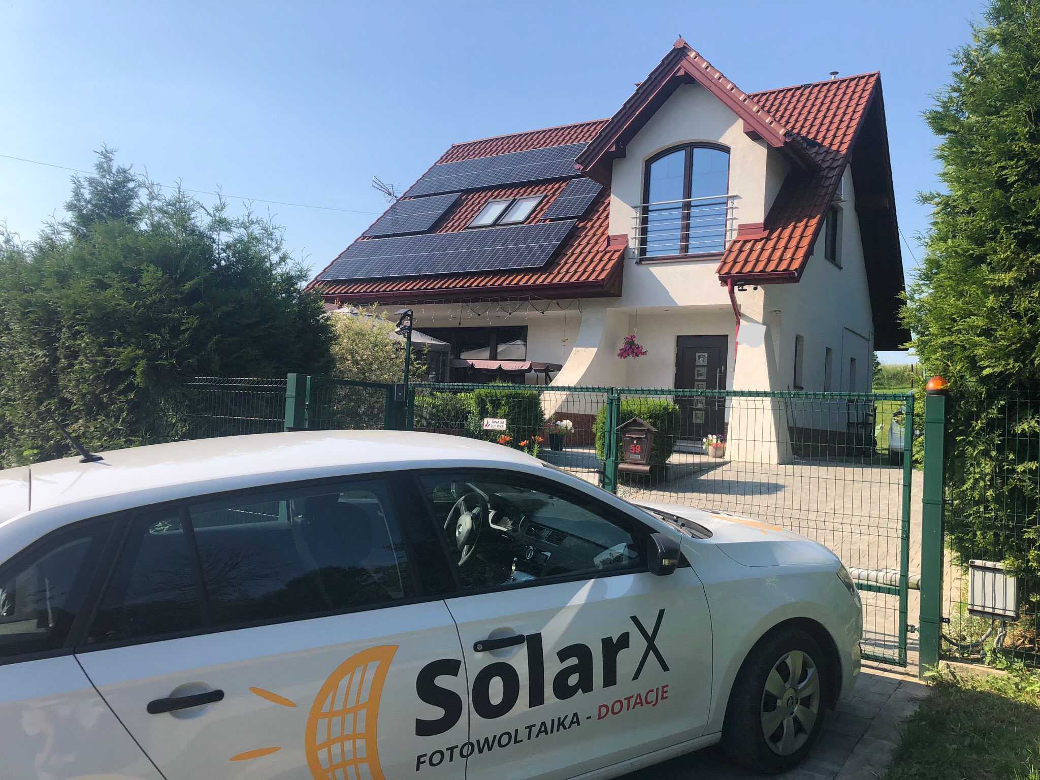 Dom jednorodzinny z panelami słonecznymi na dachu, widoczny przez bramę z zaparkowanym samochodem firmy SolarX z logo i napisem 'fotowoltaika - dotacje'.