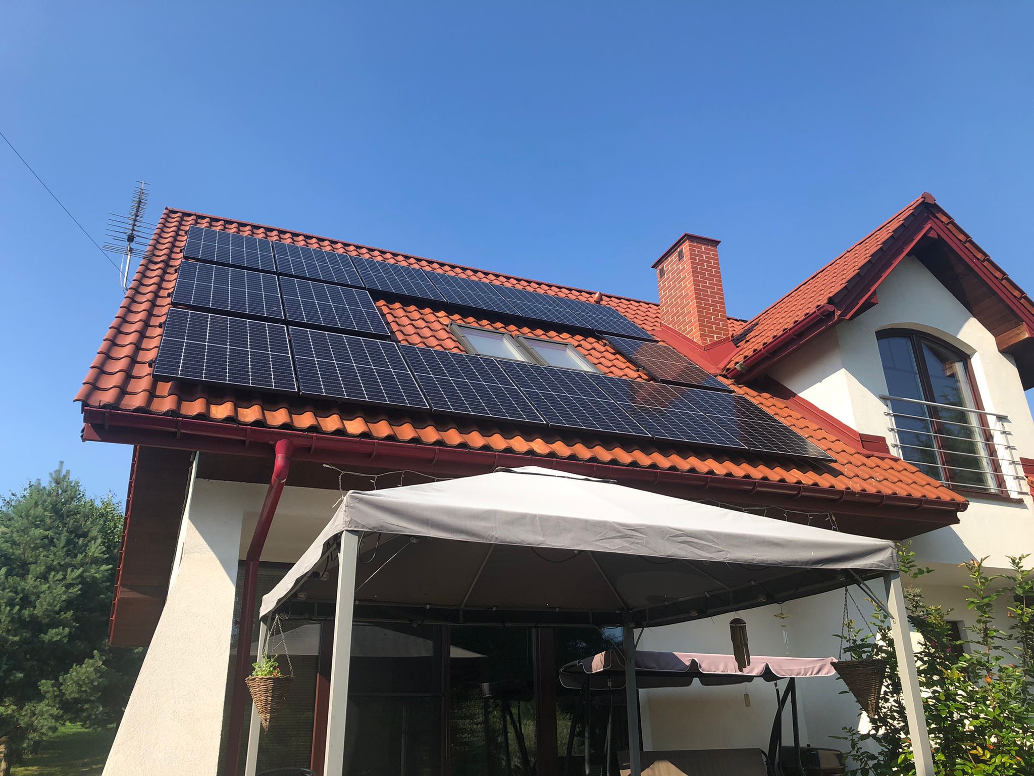 Panele słoneczne na dachu domu jednorodzinnego z czerwoną dachówką, widoczne okna dachowe, komin z cegły, antena telewizyjna oraz biała elewacja budynku z szarą altaną ogrodową w tle.