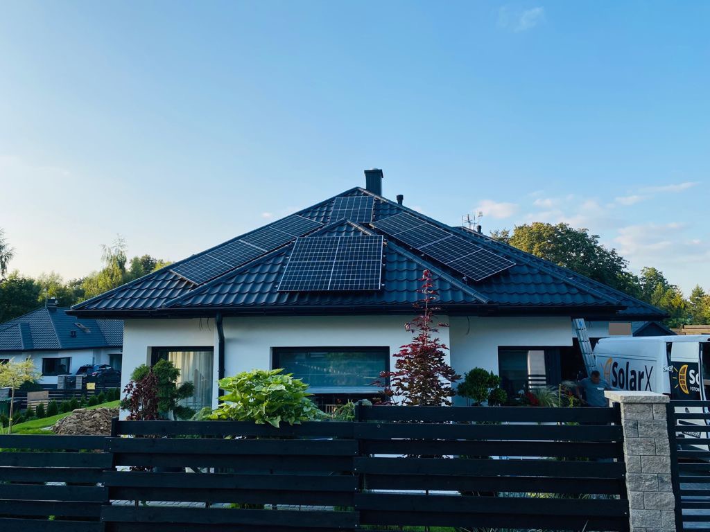 Biały dom jednorodzinny z ciemną dachówką i panelami słonecznymi na dachu, widoczny zza czarnego drewnianego ogrodzenia. Na podjeździe stoi biały samochód dostawczy z logo firmy.