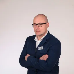 Portret mężczyzny w okularach i niebieskiej marynarce z poszetką, skrzyżowane ręce na neutralnym tle.