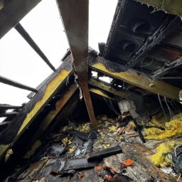 ŁUKASZ ZIELONKA CONSULTING - Spalona konstrukcja dachu z widocznymi uszkodzeniami belek, izolacji i pokrycia. Widoczne ślady pożaru i zniszczenia. Ujęcie z dołu, podkreślające skalę zniszczeń.