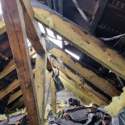 ŁUKASZ ZIELONKA CONSULTING - Spalone elementy więźby dachowej z widocznymi uszkodzeniami, fragmentami dachówek i izolacji. Widok z dołu na konstrukcję po pożarze.