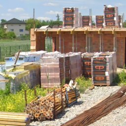 ŁUKASZ ZIELONKA CONSULTING - Budowa domu w trakcie realizacji: ściany z czerwonej cegły, materiały budowlane, betonowe podpory, zbrojenia i betoniarka na placu budowy w słoneczny dzień.