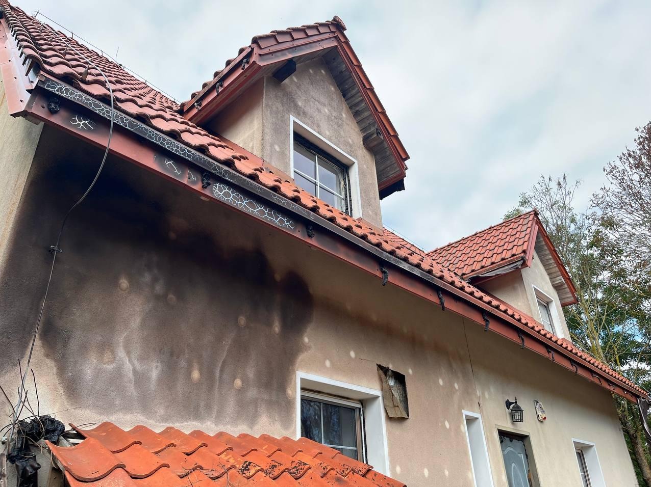 Uszkodzona elewacja domu po pożarze, widoczne osmolenia i wypalone fragmenty. Czerwona dachówka kontrastuje z ciemnymi śladami ognia na ścianie.