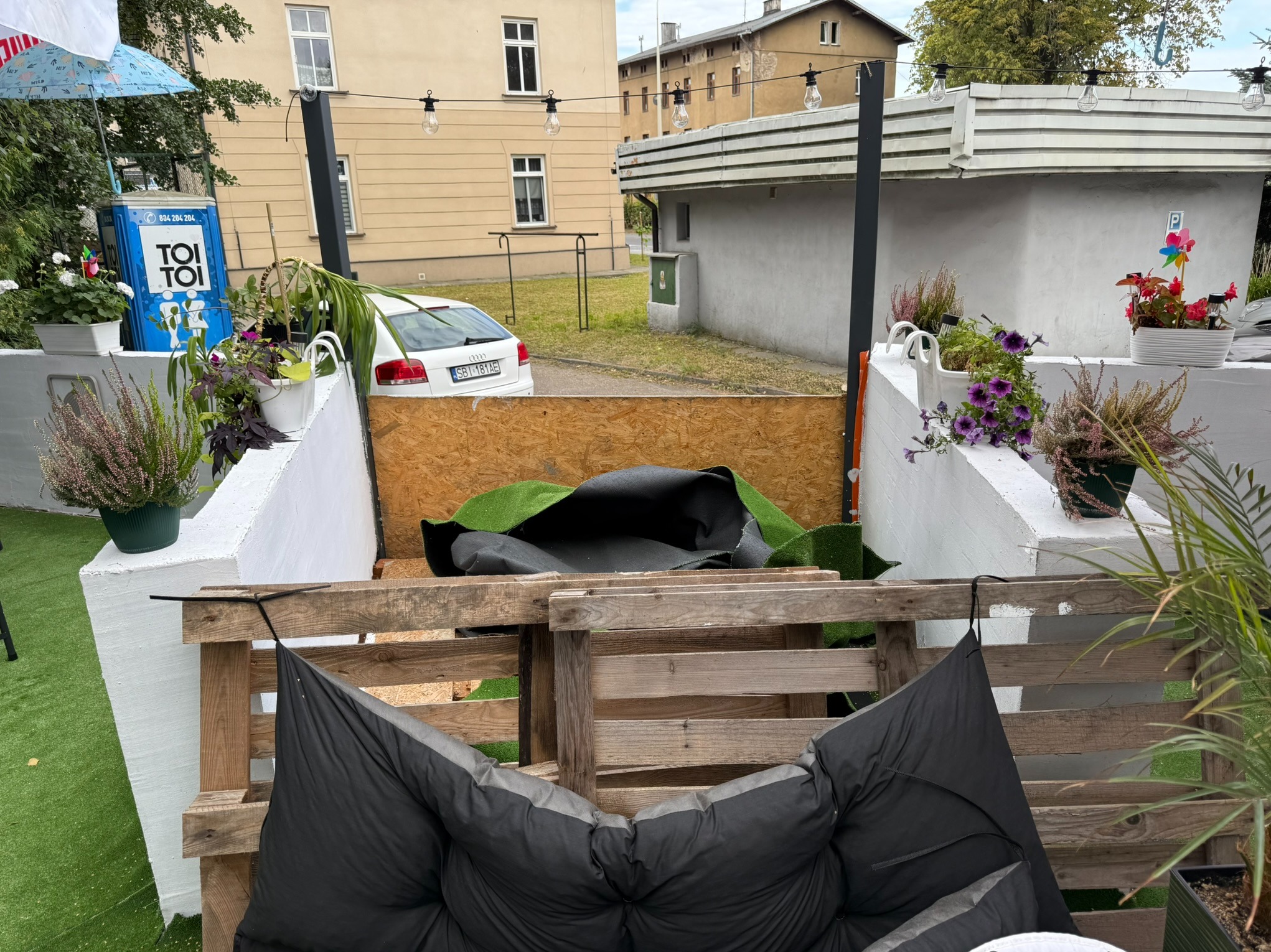 Taras z meblami z palet, dekoracjami roślinnymi i oświetleniem girlandowym. W tle budynki mieszkalne i przenośna toaleta Toi Toi.