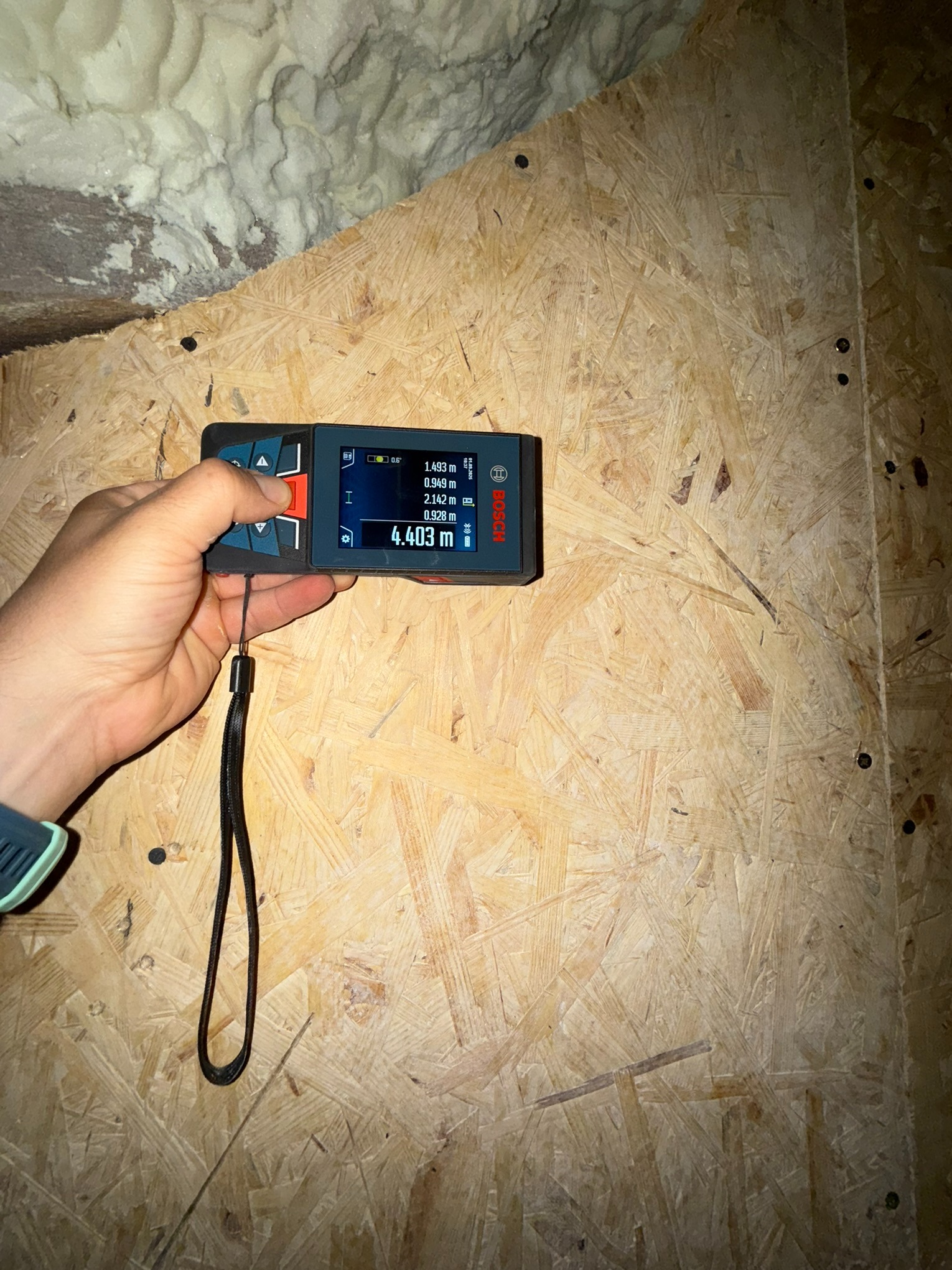 Pomiar odległości laserowym miernikiem Bosch na tle płyty OSB i pianki izolacyjnej. Wynik 4.403 m widoczny na ekranie. Kontrola wymiarów w trakcie prac budowlanych.
