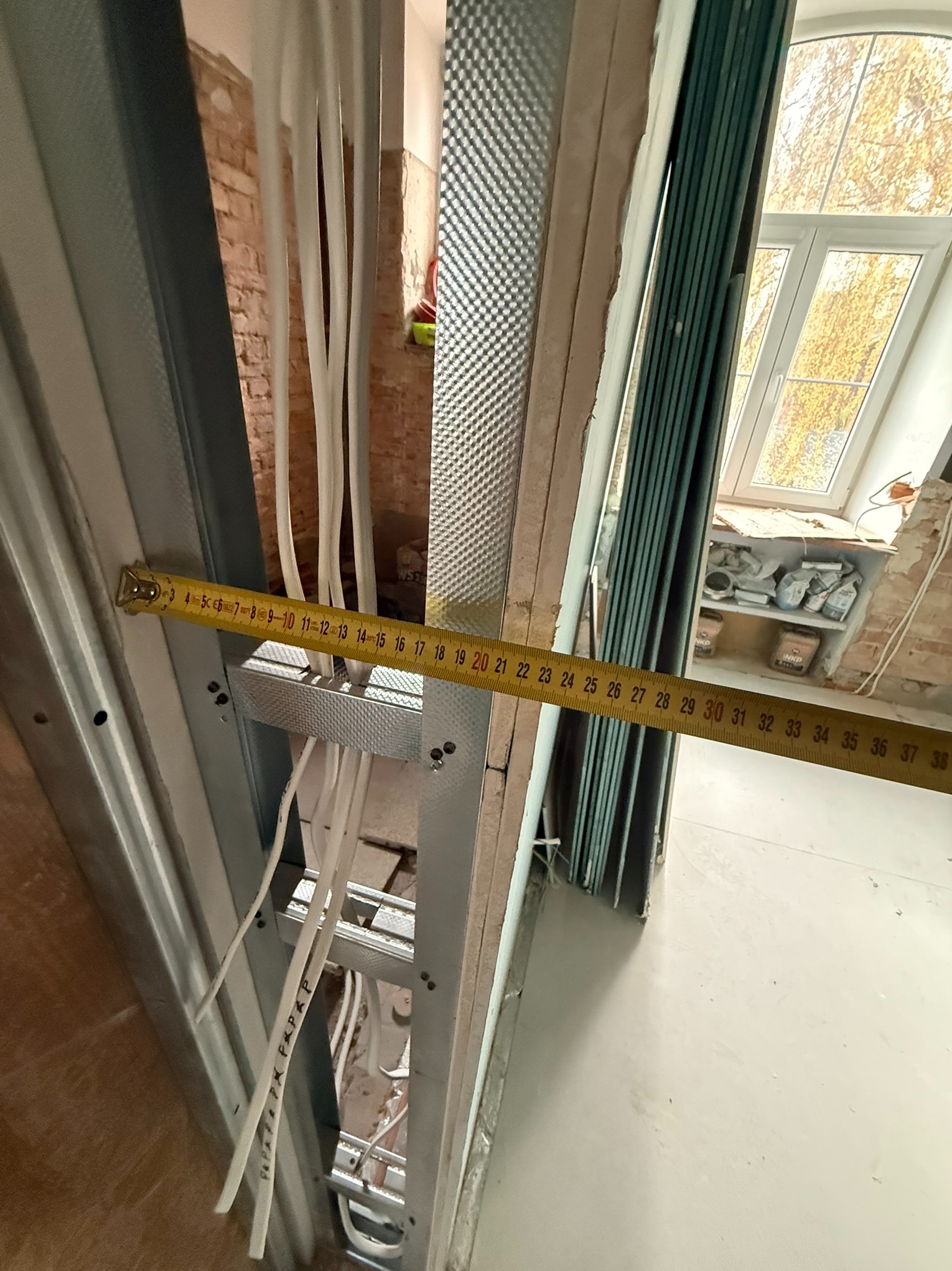 Pomiar odległości taśmą mierniczą na tle metalowej konstrukcji ściany działowej z instalacją elektryczną w trakcie remontu wnętrza z oknem na poddaszu.
