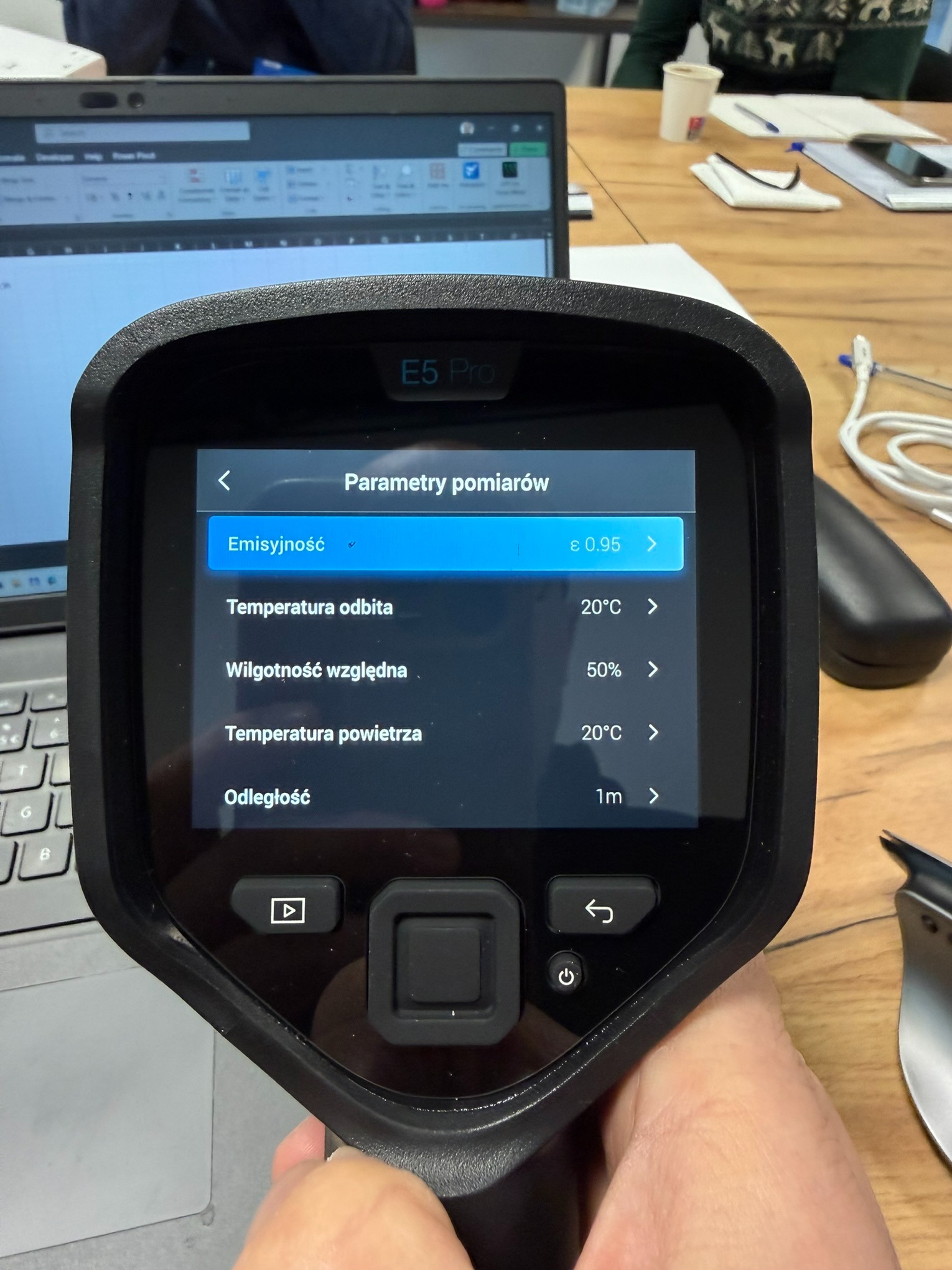Termowizor E5 Pro w dłoni, ekran pokazuje parametry pomiarów: emisyjność, temperatura odbita, wilgotność względna, temperatura powietrza i odległość.