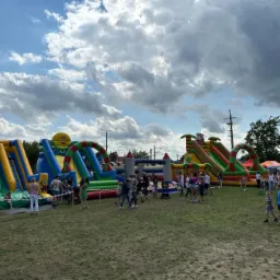 Kompleks dmuchańców z motywami Minionków i uśmiechniętej buźki, ustawiony na trawiastym terenie podczas festynu z udziałem ludzi w słoneczny dzień.