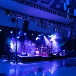 Zespół muzyczny grający na scenie oświetlonej niebieskimi światłami, widoczne instrumenty, nagłośnienie i oświetlenie estradowe.