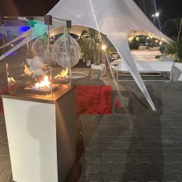 EVENT IMPERIUM - Biały namiot tipi z oświetleniem i dekoracjami, w tle palenisko gazowe w szklanej obudowie. Scena nocna, oświetlona, z elementami wystroju.