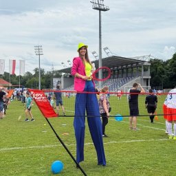 EVENT IMPERIUM - Animatorka na szczudłach w jaskrawym stroju, z rakietą tenisową, na tle boiska sportowego podczas eventu plenerowego. W tle widoczni ludzie i trybuny stadionu.