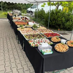 EVENT IMPERIUM - Stół cateringowy pod białym namiotem, pełen różnorodnych potraw: sałatki, przekąski, dania mięsne. W tle zieleń i biały bus. Czarny obrus, kostka brukowa.