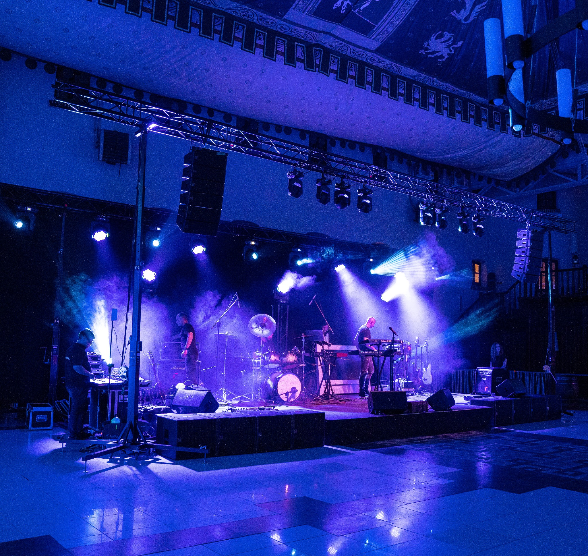 Zespół muzyczny na scenie oświetlonej niebieskimi światłami, widoczne instrumenty, nagłośnienie i efekty dymne, scena z metalową konstrukcją oświetleniową, dekoracyjne wykończenie sufitu w tle.