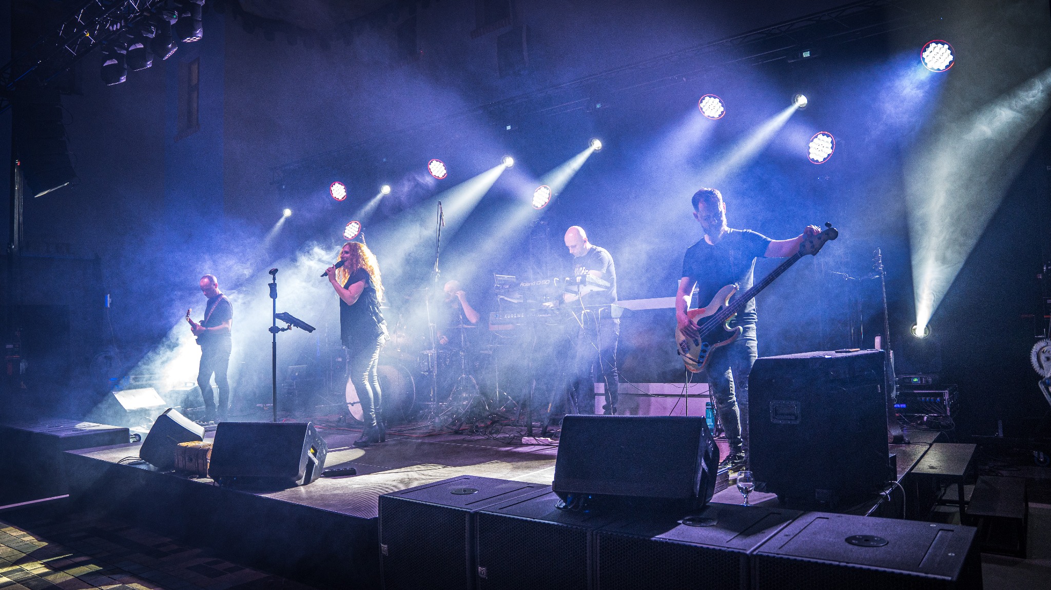 Zespół muzyczny na scenie w trakcie występu, wokalistka z mikrofonem, gitarzysta basowy i inni muzycy w oświetleniu scenicznym z efektem dymu.