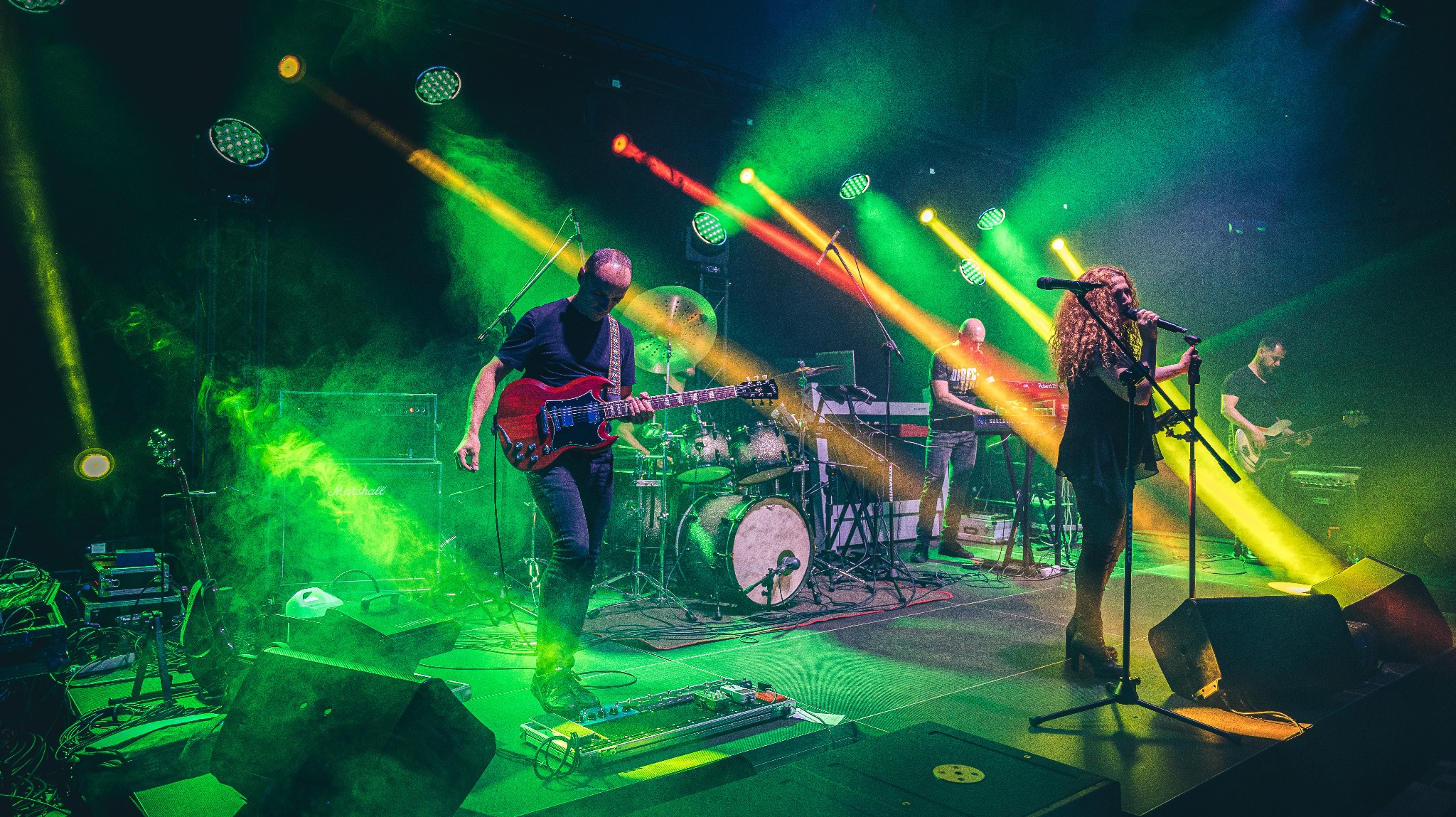 Zespół muzyczny na scenie podczas koncertu oświetlonego zielonymi i żółtymi reflektorami, wokalistka z kręconymi włosami śpiewa do mikrofonu, gitarzysta gra na czerwonej gitarze elektrycznej...