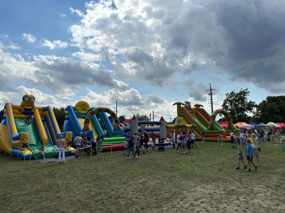 Kompleks dmuchańców z motywami Minionków i uśmiechniętej buźki, ustawiony na trawiastym terenie podczas festynu z udziałem ludzi w słoneczny dzień.