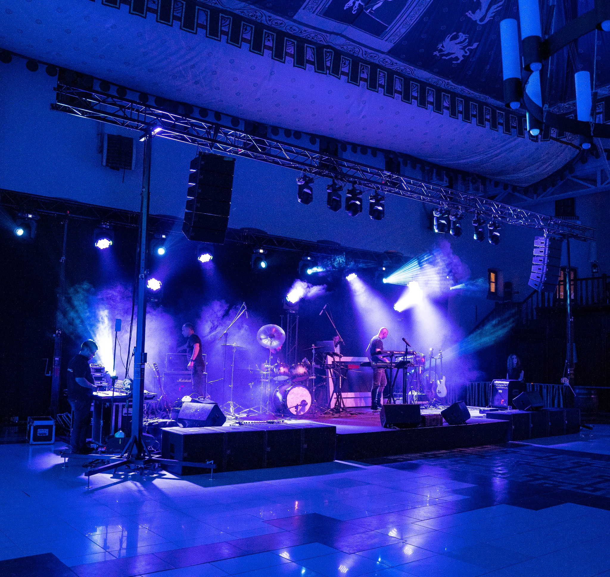 Zespół muzyczny grający na scenie oświetlonej niebieskimi światłami, widoczne instrumenty, nagłośnienie i oświetlenie estradowe.