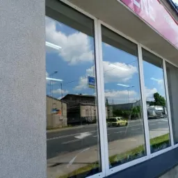 Duże, czyste okna wystawowe z białymi ramami, odbijające błękitne niebo z chmurami i uliczny ruch, widoczne budynki po drugiej stronie ulicy.