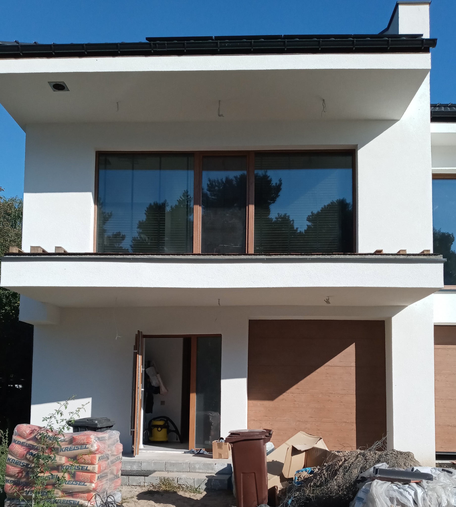 Nowoczesny dom w trakcie porządkowania po budowie: worki z zaprawą, śmieci i odkurzacz budowlany przed wejściem.